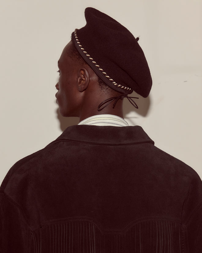 A person in a dark jacket is seen from behind, wearing the Script Beret in black with brown/black piping, made of 100% wool in France, set against a light background.