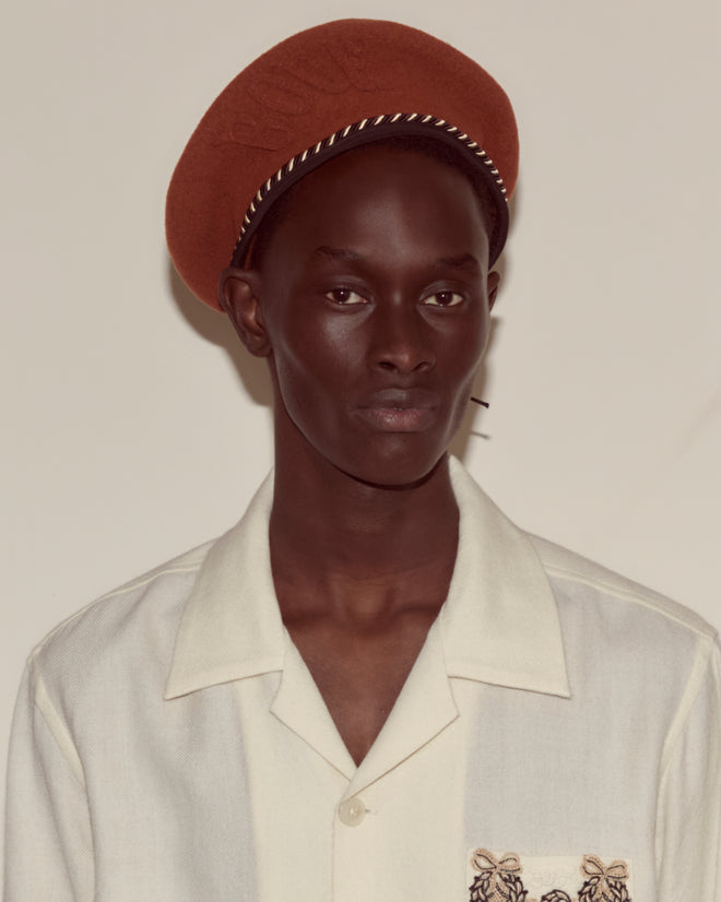 Person wearing a cream shirt with an embroidered pocket and the Script Beret in Rust with Brown/Black Piping, 100% wool, made in France, against a plain background.