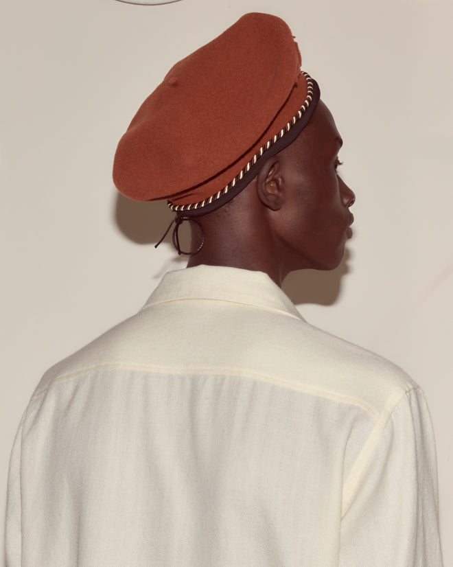 Person in a white shirt wears the Script Beret—Rust with Brown/Black Piping, shown from behind against a plain background.