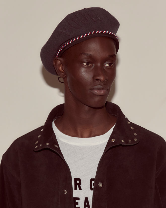 A person wearing the Script Beret in charcoal with red/blue piping and a jacket with metal studs over a white printed shirt, looking to the side.
