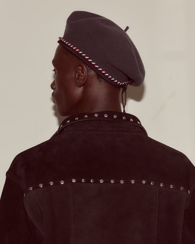 Person in a dark studded jacket and Script Beret - Charcoal with Red/Blue Piping, shown from the back against a plain background.