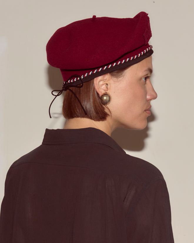Woman in a Script Beret - Bordeaux with Red/Blue Piping, worn from the side with a dark shirt against a plain background. This classic beret style is made in France.