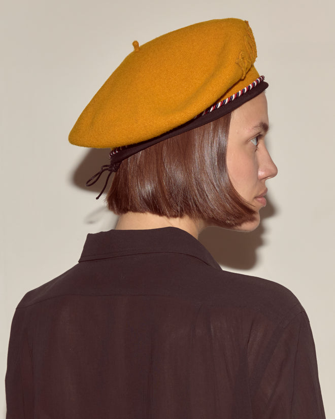 Woman in a Script Beret - Gold with Red/Blue Piping and a dark shirt faces sideways against a plain background.