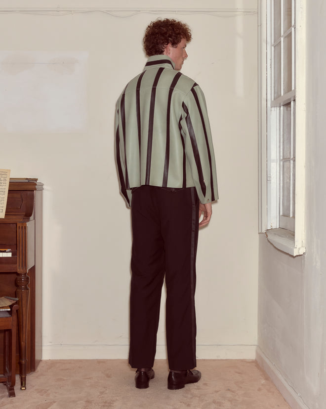 A man in a Seafoam Stripe Welding Jacket - Seafoam/Black and black pants stands facing a wall near a window and a piano.