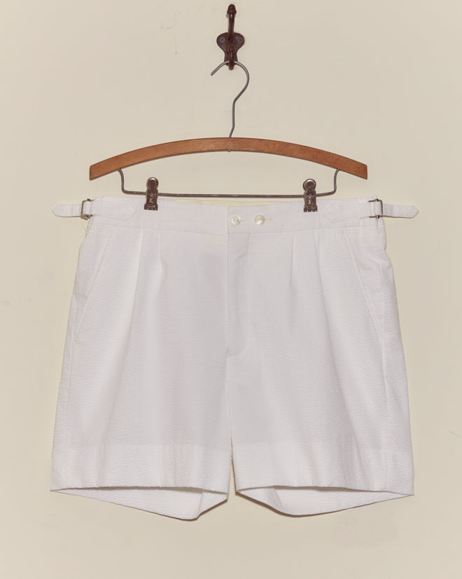 Seersucker Double-Button Shorts hang on a wooden hanger against an off-white wall, showcasing their crisp pleated design with side adjusters for a vintage style look.