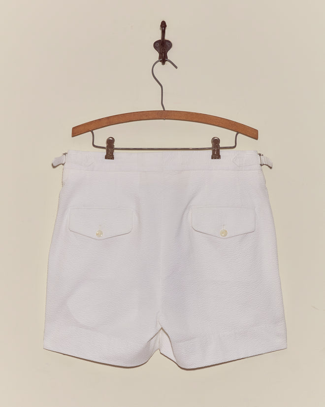 The Seersucker Double-Button Shorts, featuring white pleats and two back pockets, hang on a wooden hanger against a plain wall—ideal for bringing vintage style to your summer outfit.