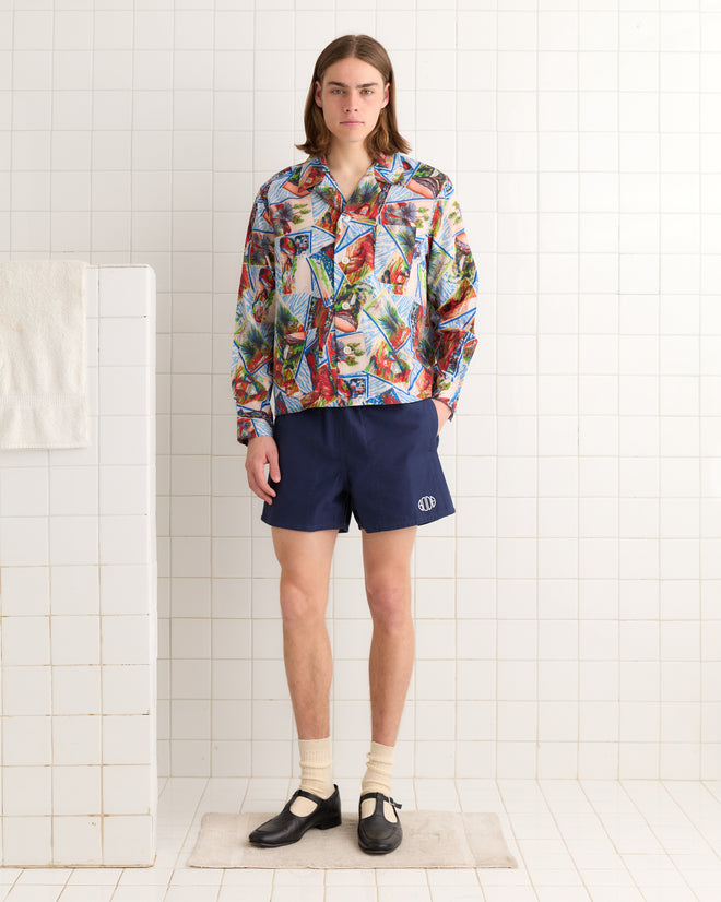 A person in a colorful Seersucker Postcard Long Sleeve Shirt inspired by 1950s fabric, navy shorts, socks, and black shoes stands in a tiled room.
