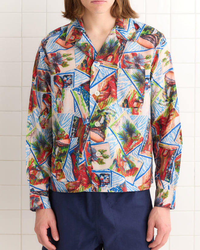 Wearing a Seersucker Postcard Long Sleeve Shirt, designed with a vibrant 1950s tropical pattern, this individual stands against a tiled wall, capturing the essence of vintage tropical postcards.