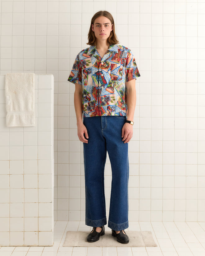 Dressed in a vibrant Seersucker Postcard Short Sleeve Shirt and jeans, an individual stands in a tiled room with a towel hanging over the partition wall.