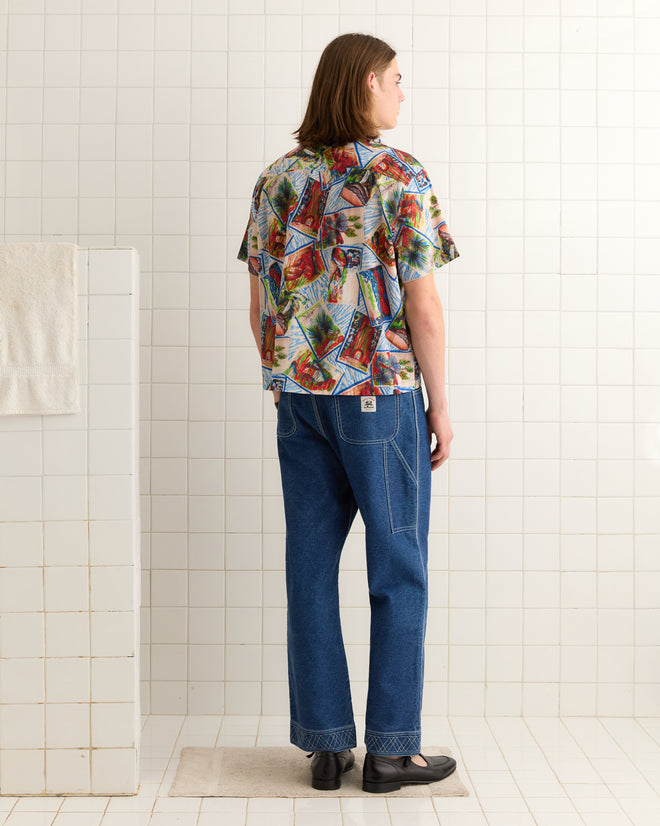 A person with long hair, wearing a colorful printed Seersucker Postcard Short Sleeve Shirt and jeans, stands in a tiled room facing away.