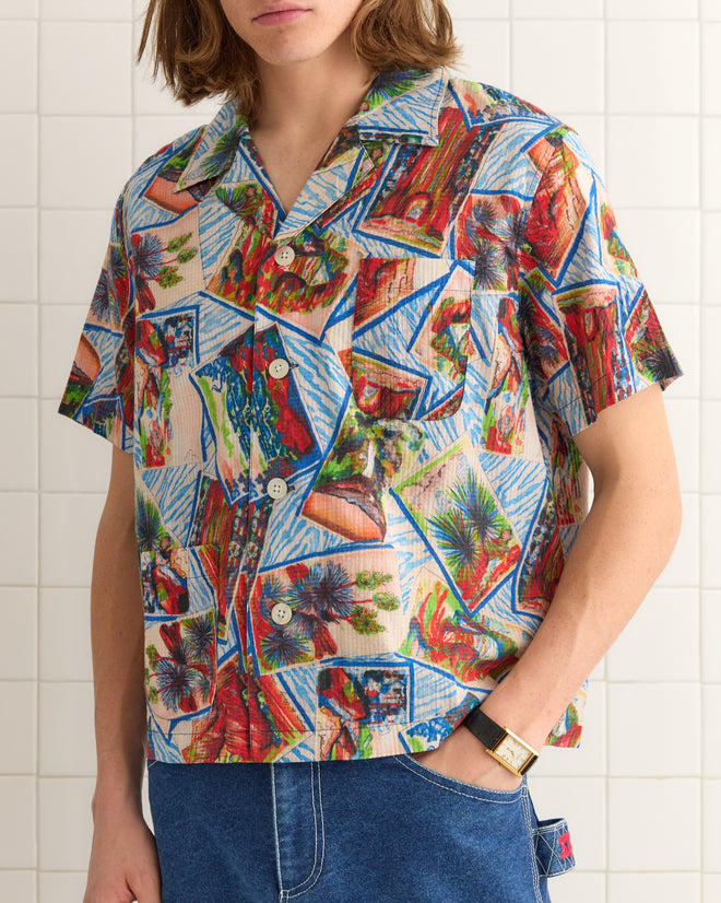 Clad in a Seersucker Postcard Short Sleeve Shirt and blue jeans, a person stands against a tiled wall, effortlessly blending style and comfort.