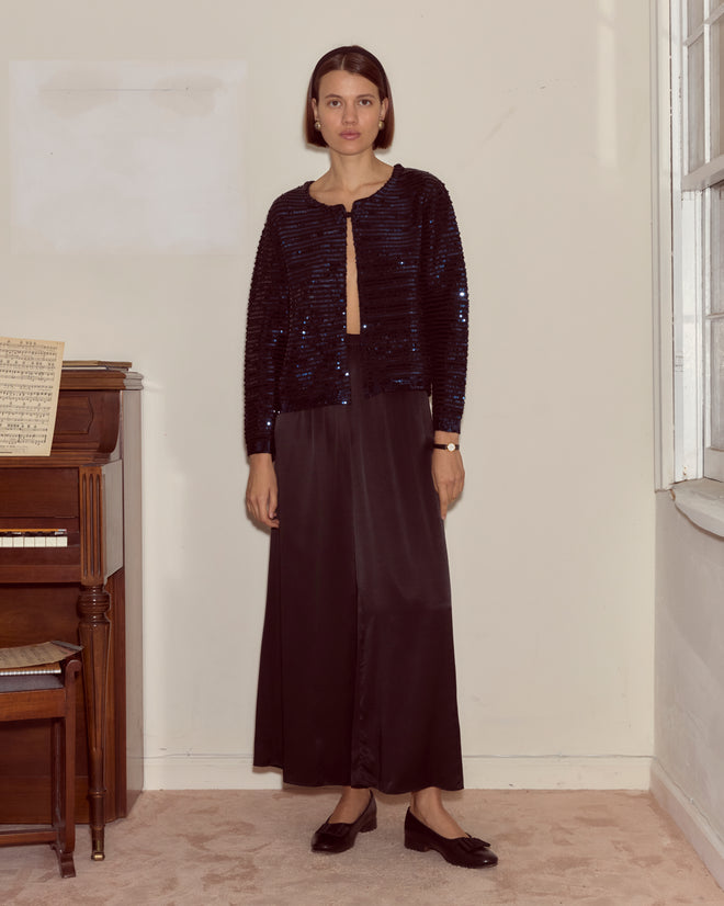 A woman wearing the Sequin Spotlight Swing Cardigan in black/blue stands indoors beside a piano and a window.