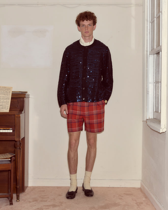 A man in red plaid shorts, the Sequin Spotlight Cardigan - Black, and loafers stands indoors beside a piano.