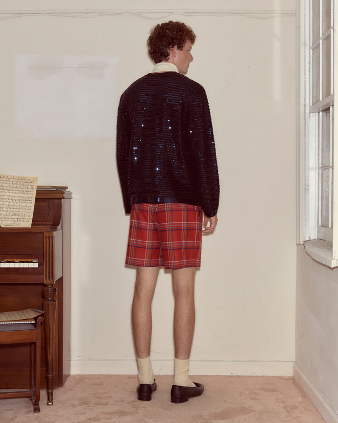 A person stands facing a wall, wearing the Sequin Spotlight Cardigan - Black and red plaid shorts.