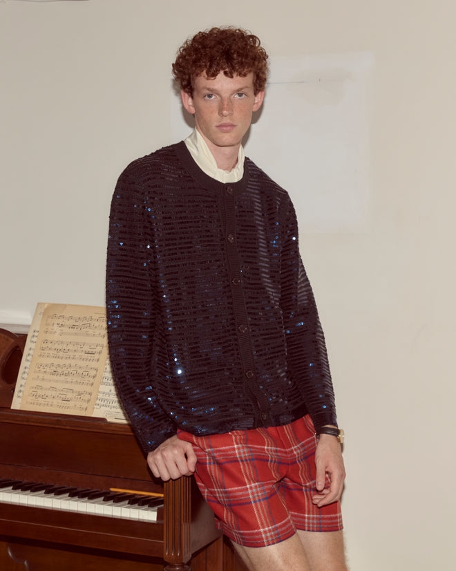 A man stands by a piano with sheet music, wearing the Sequin Spotlight Cardigan - Black and red plaid shorts, making a statement with his bold knitwear.