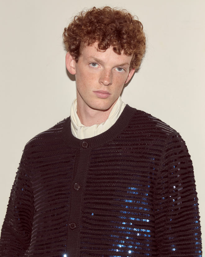 A person with curly red hair stands against a plain wall, wearing the Sequin Spotlight Cardigan in black—an elegant knitwear piece—layered over a cream shirt.