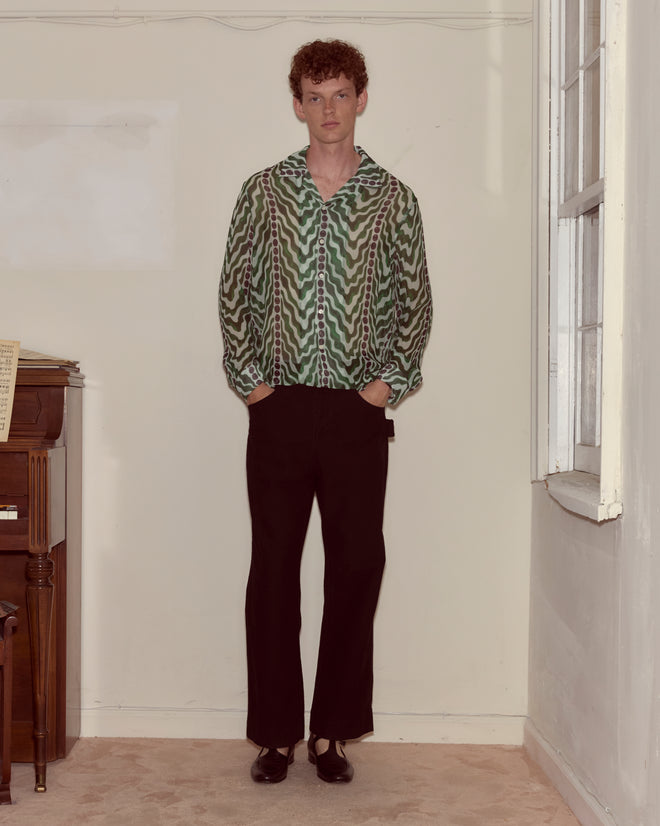 A person stands indoors by a window and piano, hands in pockets, wearing the Serpentine Long Sleeve—a green silk crepe shirt with a 1930s pattern—and black pants.