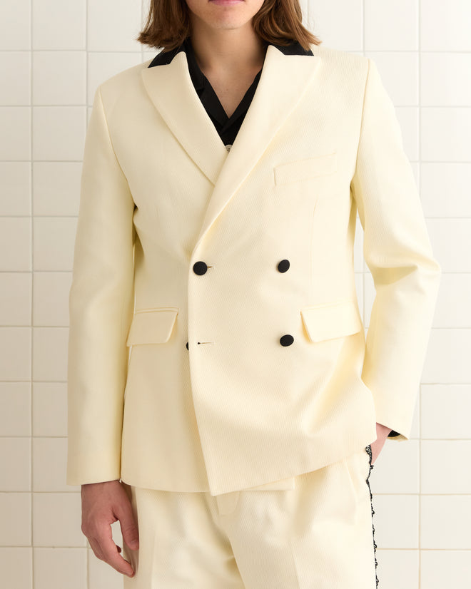 A person confidently stands against a tiled wall in a Seymour Double-Breasted Suit Jacket, crafted from heavyweight cream twill and featuring striking black buttons.