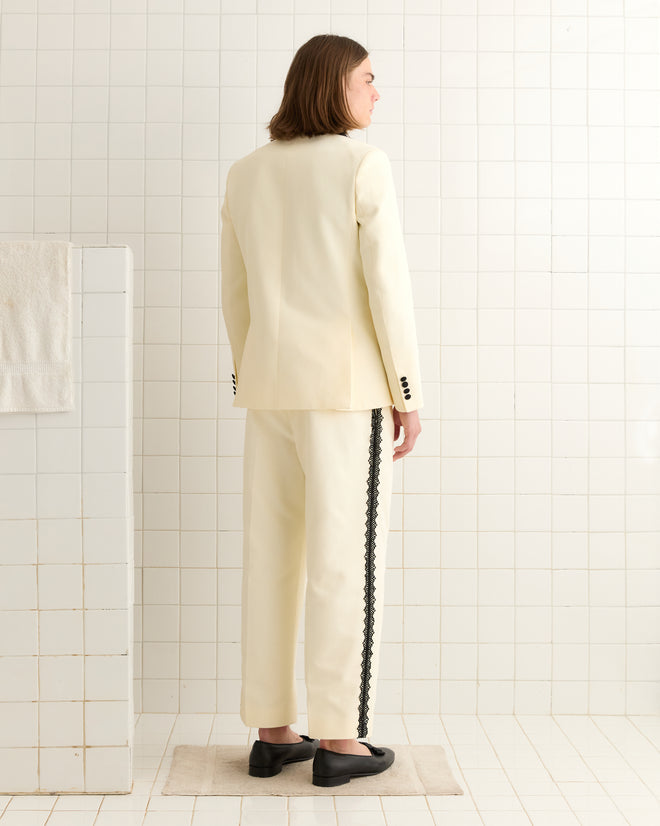 Someone in a Seymour Double-Breasted Suit Jacket, cream with black trim, stands on a mat in a tiled room, facing away.