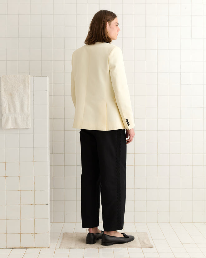 A person with long hair in a cream blazer and Seymour Suiting Trousers - Black stands in a white tiled room, facing away.