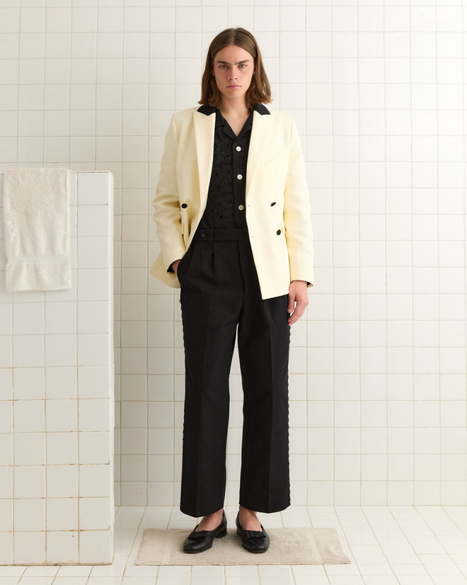 A person in a white cotton twill double-breasted blazer and Seymour Suiting Trousers in black with scalloped lace details, standing on a tiled floor.