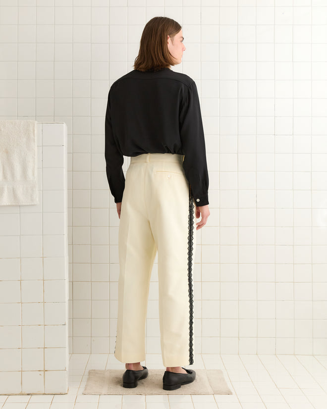 A person wearing a black shirt and Seymour Suiting Trousers in white stands in a tiled room, facing away.