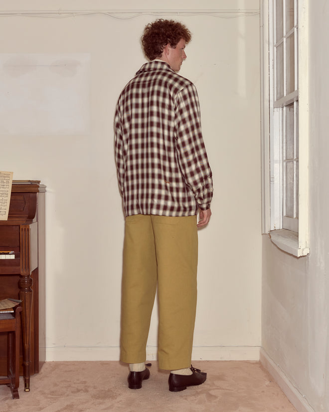 A person with curly hair, wearing a Shadow Plaid Franck Jacket and khaki pants, stands facing a wall in a sparsely furnished room.