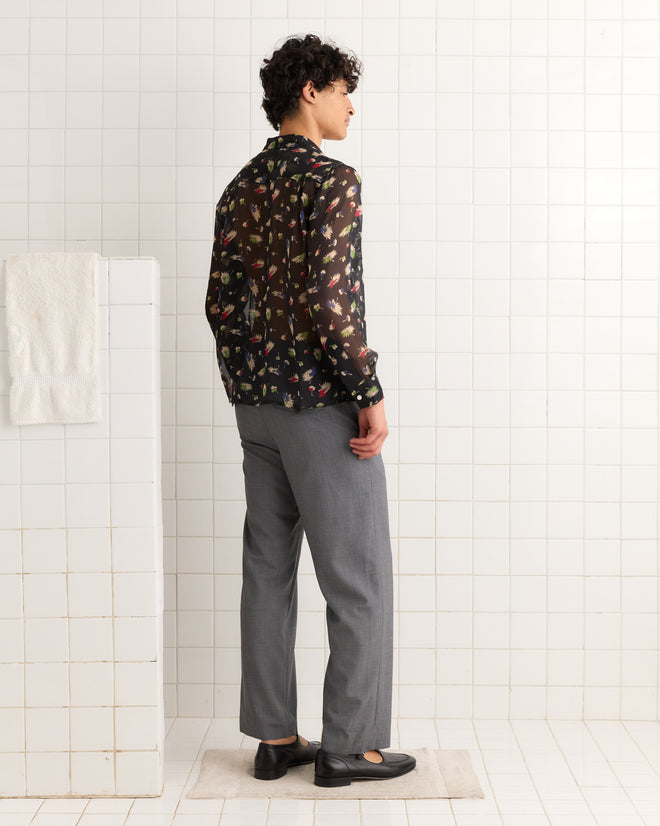 Person standing in a tiled room, facing sideways, wearing gray pants and the Sheer Midnight Daisy Shirt—an elegant black, sheer, floral print silk top made in India.