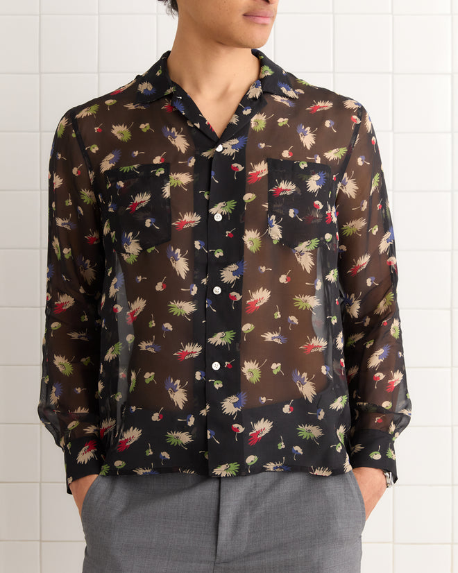 Someone wearing the Sheer Midnight Daisy Shirt—a black, sheer silk button-up with a floral print and chest pockets—paired with gray pants.