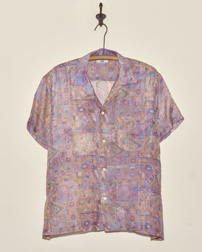 The Sheer Puzzle Shirt (M/L), a short-sleeved vintage top with a unique pattern in pastel shades, hangs on a wooden hanger against a plain wall.