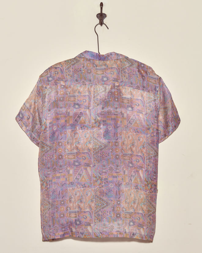 The Sheer Puzzle Shirt (M/L) features pastel patterns and short sleeves, displayed on a wall hook with its back facing forward.