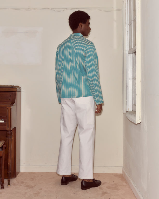 Wearing the Shore Stripe Long Sleeve Shirt with blue and white stripes and white pants, a person stands indoors facing a window, back to the camera.