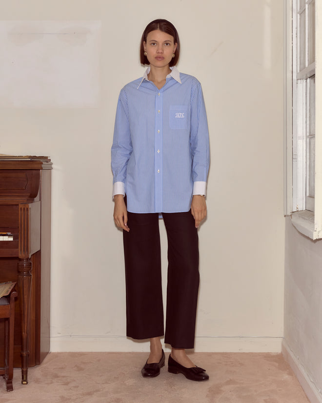 A woman stands indoors next to a piano wearing a blue Signet Murray Shirt, black pants, and black shoes.