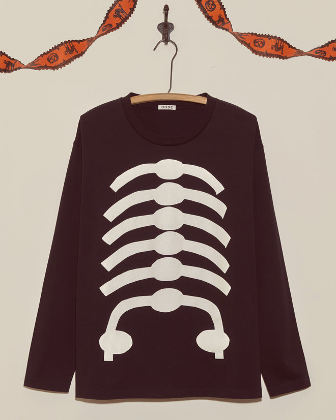 The Skeleton Long Sleeve Tee is a black shirt with a white rib cage print, hanging on a hook beneath an orange Halloween garland—a vintage Halloween classic perfect for spooky season.