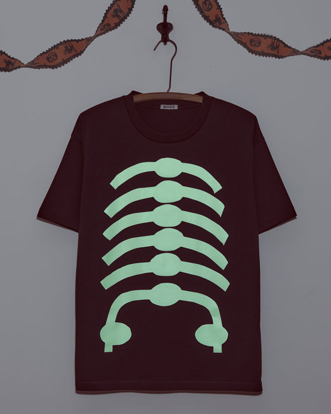 The Skeleton Tee, a black glow-in-the-dark shirt with a bold green ribcage design, hangs on a hanger against a gray wall.