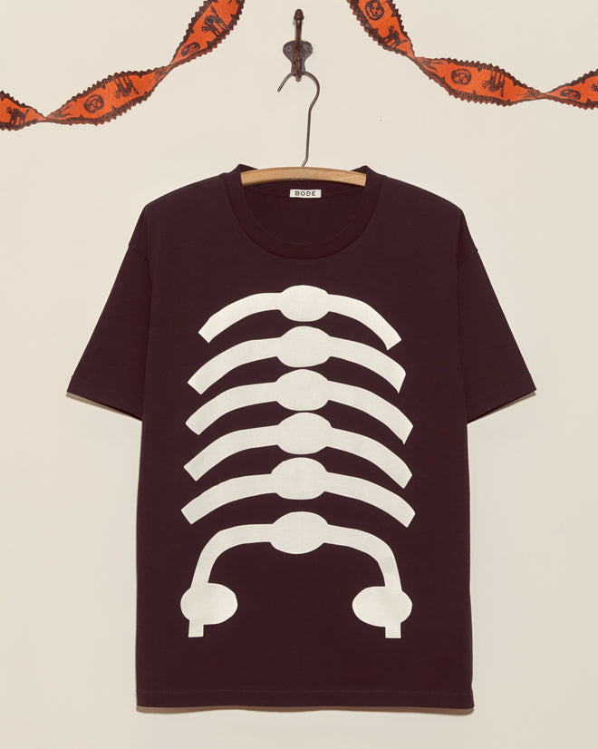 The Skeleton Tee, a black shirt with a white ribcage design, hangs on the wall with orange Halloween garland above—a vintage-style top ideal for fans of unique Halloween shirts.