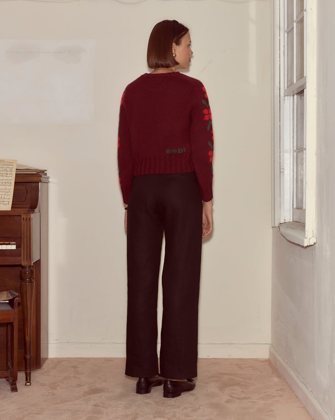 A woman with short brown hair stands facing away, wearing a maroon Skunk Sweater and black pants in a room with a piano.