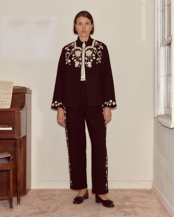 A woman in black pants and a Snowdrop Appliqué Jacket stands by a piano in a bright room.
