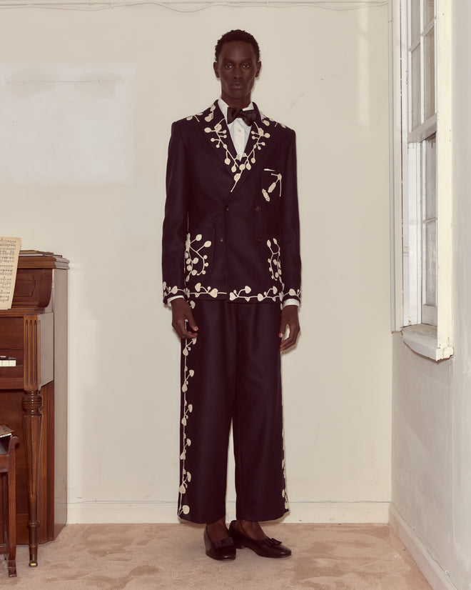 A person in the Snowdrop Appliqué Suit Jacket in navy, featuring white floral appliqué, stands indoors near a piano and a window.