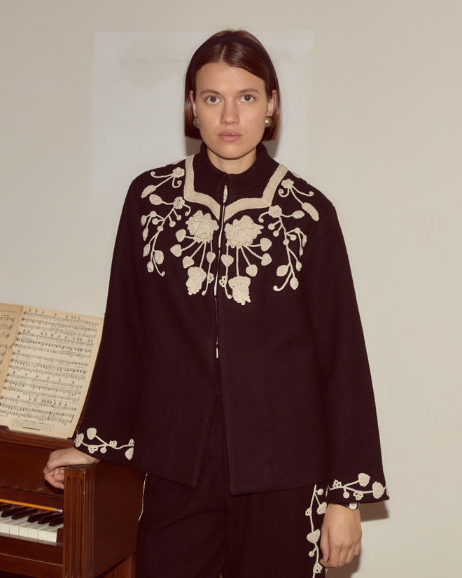 A woman in the Snowdrop Appliqué Jacket stands by a piano with sheet music, the elegant floral embroidery enhancing the charm of her swing silhouette wool outerwear.