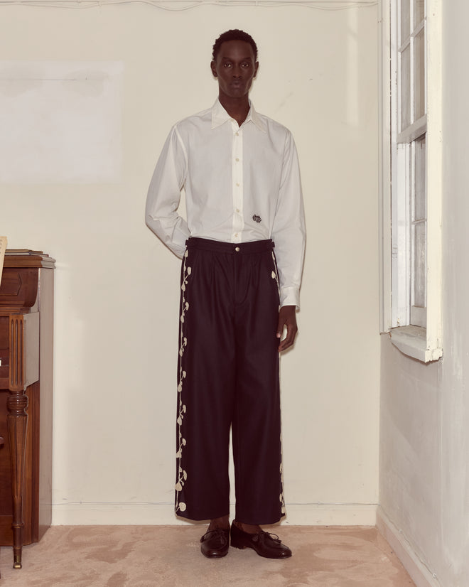 A person stands indoors wearing a white shirt and Snowdrop Appliqué Trousers in navy, featuring side embroidery.