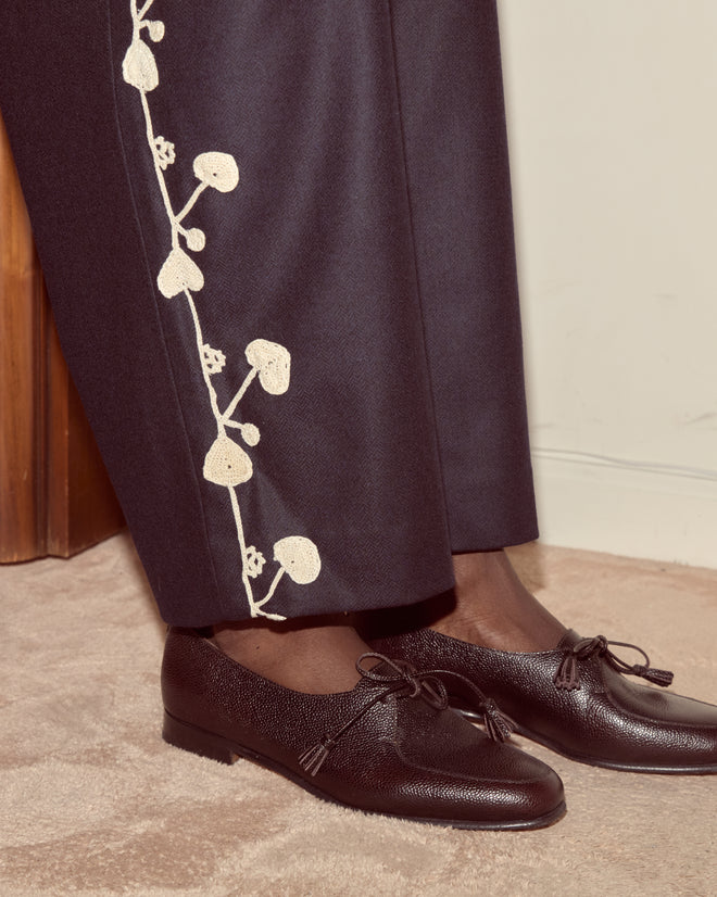 Person wearing Snowdrop Appliqué Trousers in navy—dark wool suiting with hand-crocheted white mushroom details—styled with black leather shoes, standing on a beige carpet.