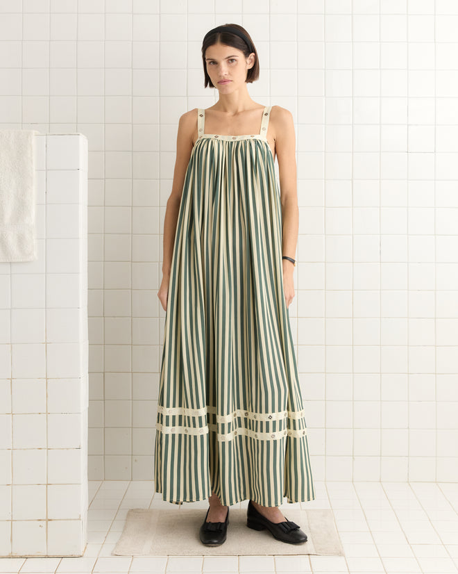 Wearing the Soda Stripe Dress, a person in a 1940s-inspired, long grey-green striped design stands in a tiled room beside a towel on a partition.