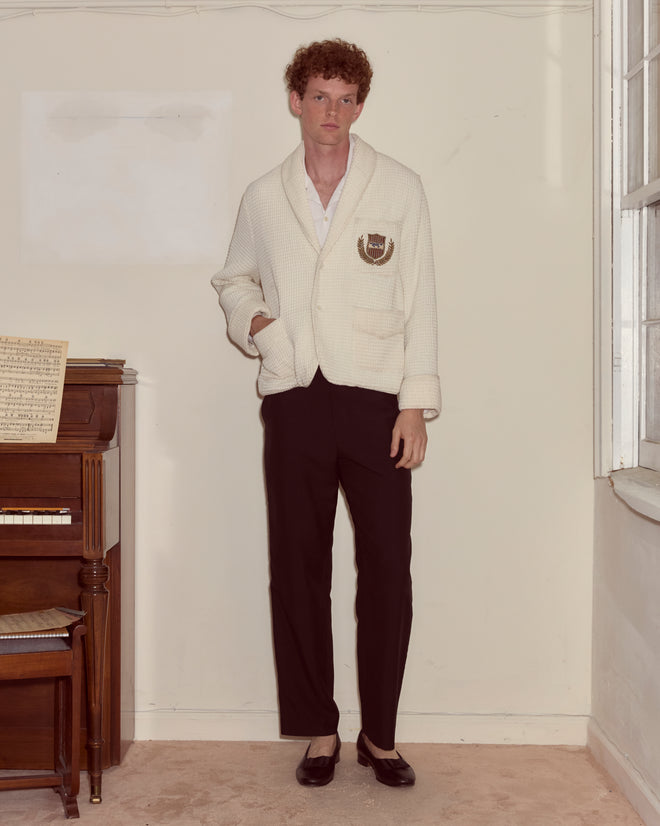 A man wearing a Sportsman Jacket and black pants stands by a piano in a softly lit room.