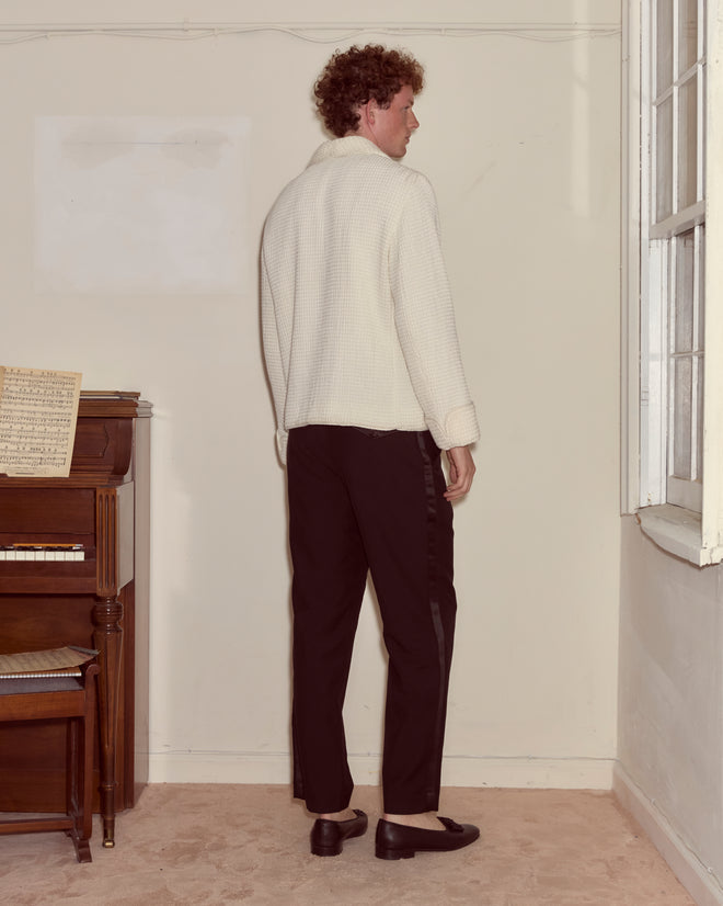 A person wearing a Sportsman Jacket and black pants stands facing away next to a piano and a window in a beige room.