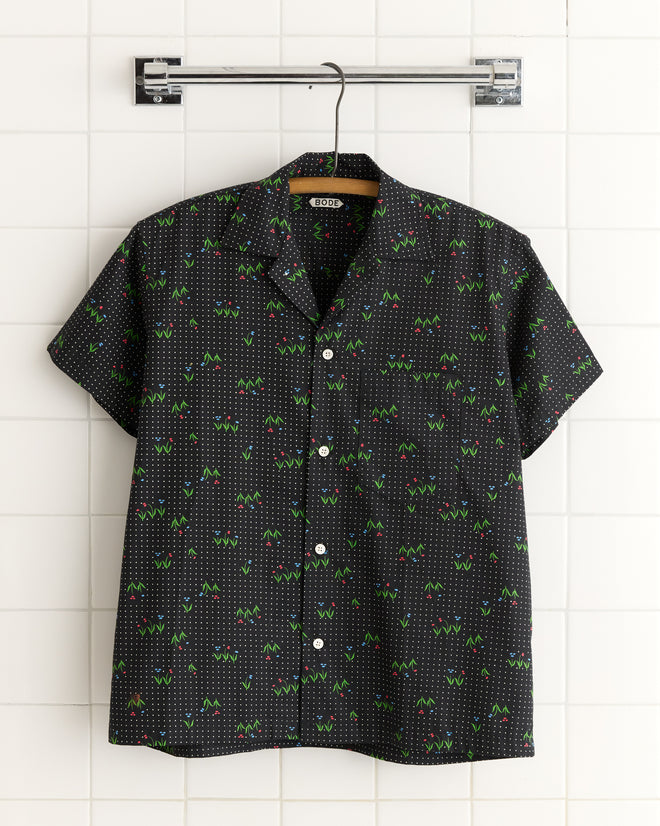 The Spring Grass Short Sleeve Shirt - XS/S, a black button-up with colorful geometric patterns made from deadstock fabric, hangs on a hanger against a tiled wall.