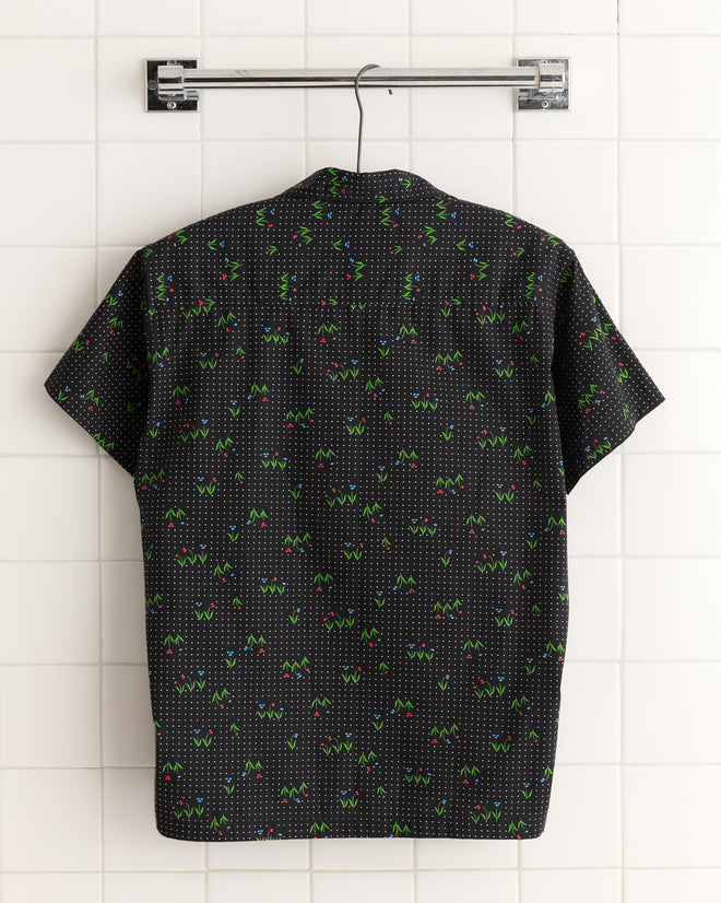 The Spring Grass Short Sleeve Shirt - XS/S features a colorful geometric pattern inspired by budding flowers. Displayed on a metal hanger against white tiles, this unique piece is perfect for enhancing any wardrobe.