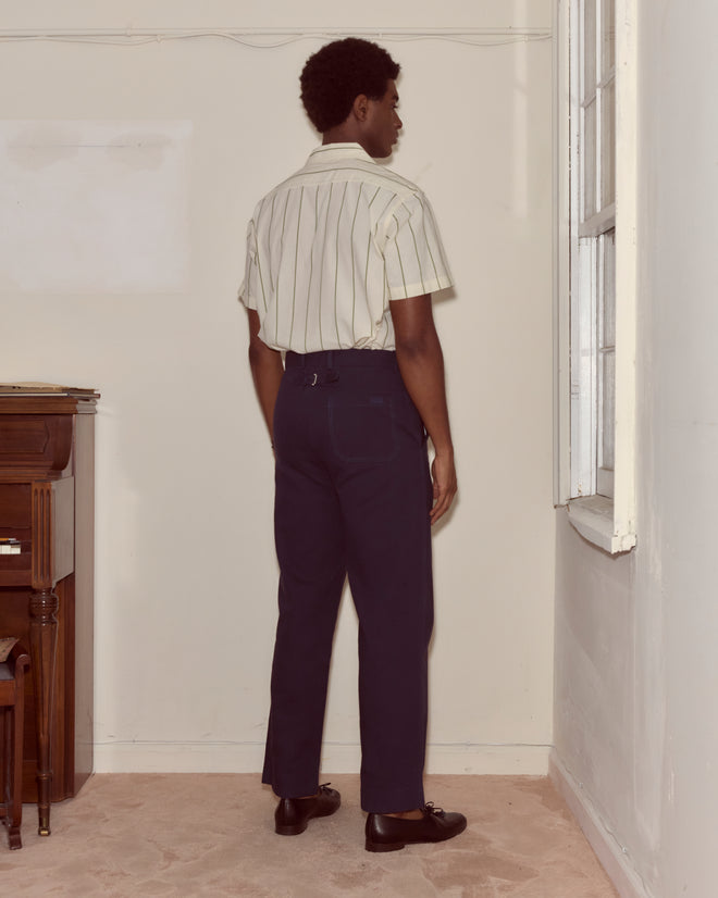 A person in a striped shirt and Standard Trousers - Navy stands facing the corner of a room near a window.
