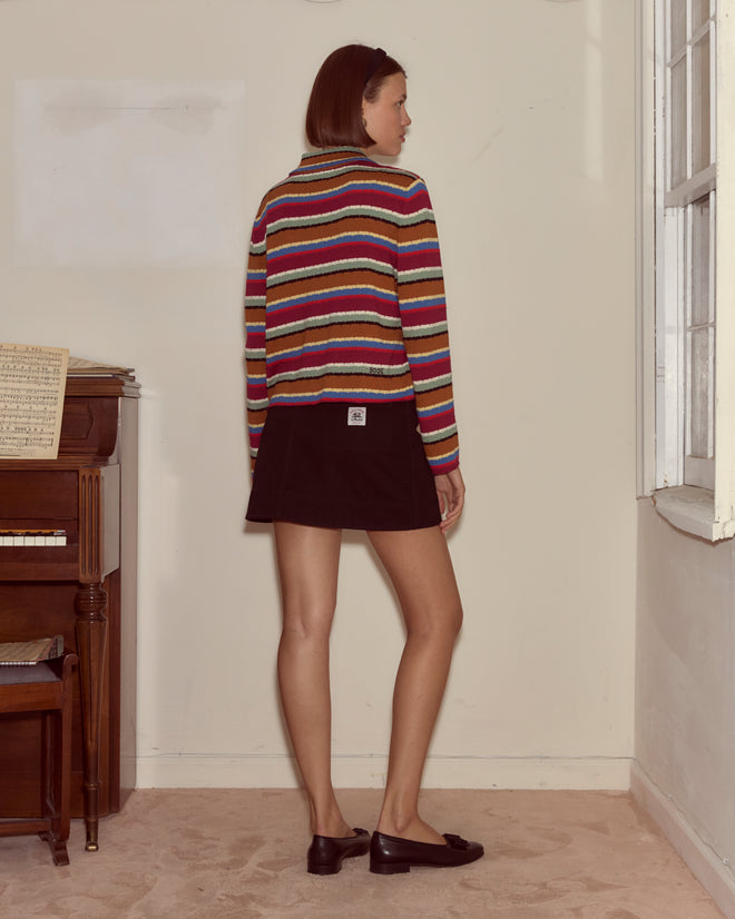 A woman wearing the Starkey Cardigan and a black skirt stands near a window, facing away in a carpeted room with a piano.
