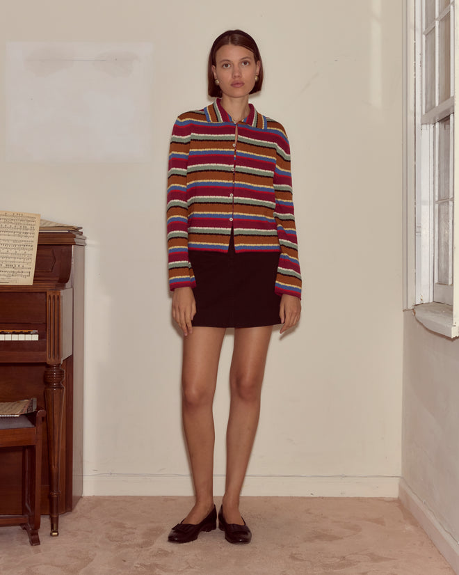 A woman in a Starkey Cardigan and black skirt stands indoors beside a piano and a window.